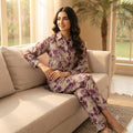 A woman wearing a matching floral pajama set sits on a beige sofa in a bright living room. The set includes a long-sleeve button-up shirt and coordinating pants, featuring a purple and green floral pattern on a light beige background.