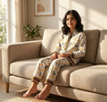 A young girl sits barefoot on a beige fabric sofa in a sunlit living room, wearing a cream pajama set with a yellow and gray floral print and brown decorative trim.