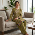 A woman sits on a beige fabric sofa wearing an olive green traditional outfit with intricate multicolor embroidered patterns and a geometric border. The setting is a bright, modern living room with a window, plants, and a wooden coffee table.