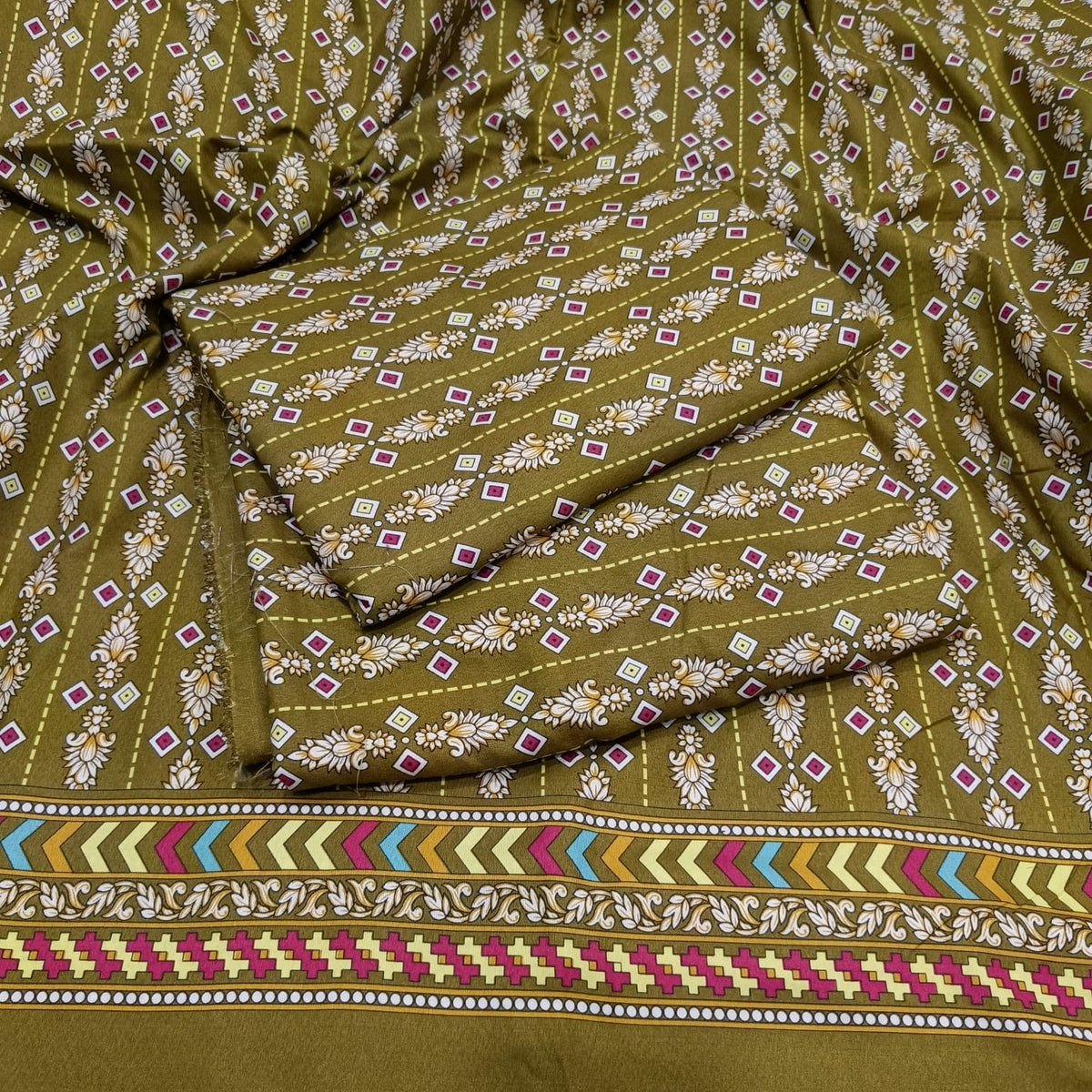 An olive green fabric with a detailed block print pattern of white flowers, small pink and green diamonds, and a colorful geometric border at the bottom.