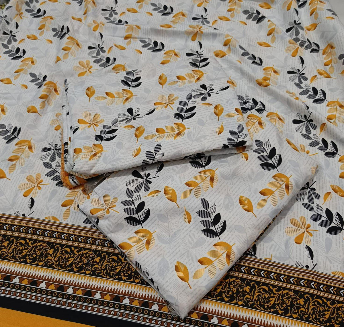 A folded piece of white fabric with a repeating pattern of mustard yellow and dark gray leaf motifs, featuring a decorative black and gold border along the bottom edge.