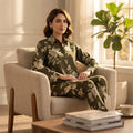 A woman sits in a beige armchair in a sunlit room, wearing a matching long-sleeve button-up shirt and pants set in olive green with a beige floral and leaf pattern.