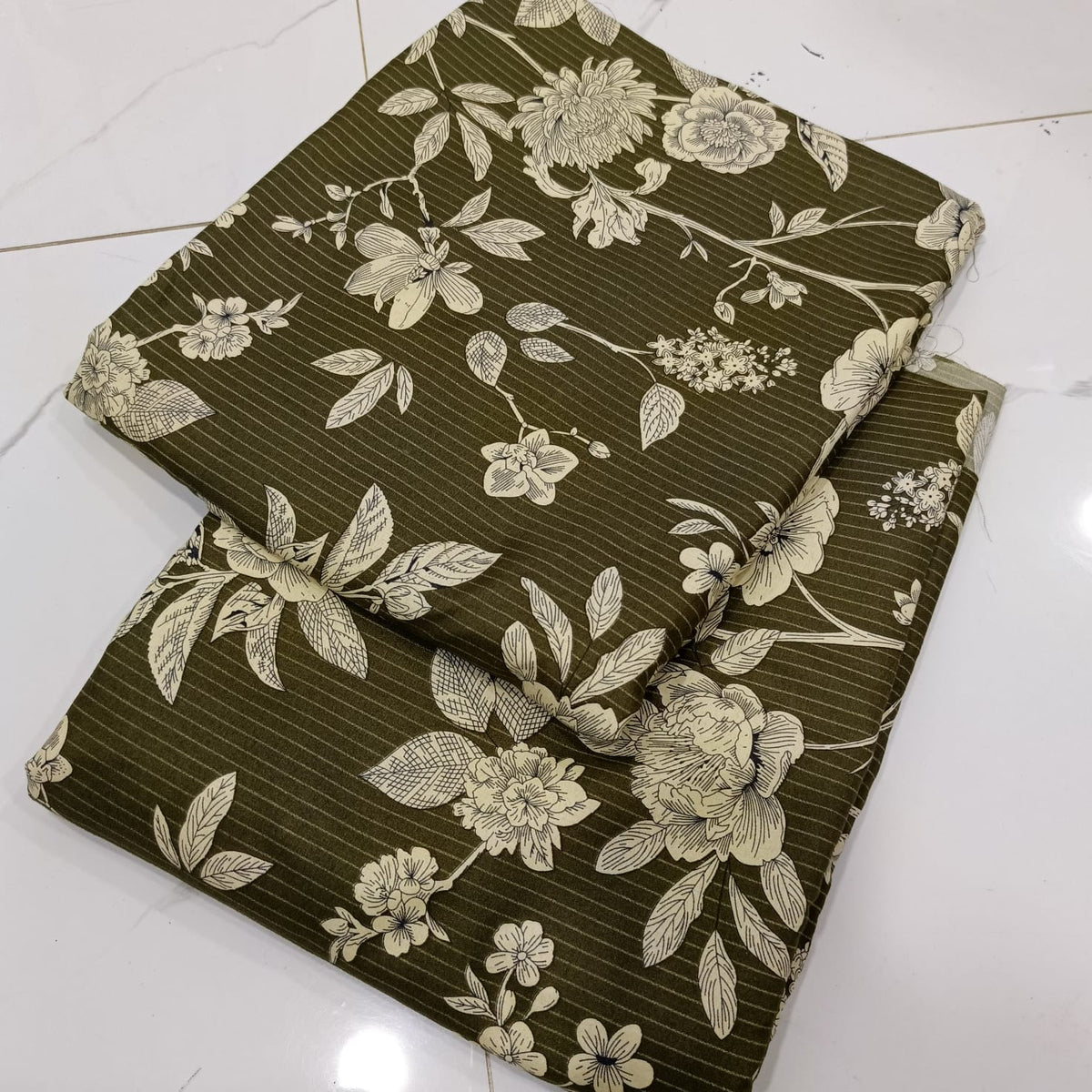 A folded piece of olive green fabric with a beige floral and leaf pattern, laid on a white tiled floor.
