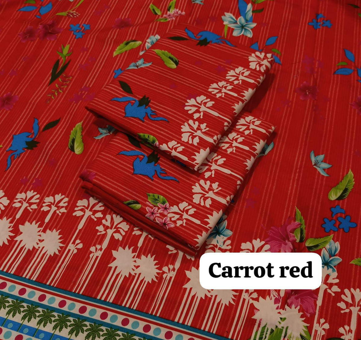 A folded fabric set in a bright carrot red color with a striped and floral print, displayed on a matching red background. The set includes a larger cloth and two smaller folded cloths, decorated with white flowers, blue birds, and green leaves.